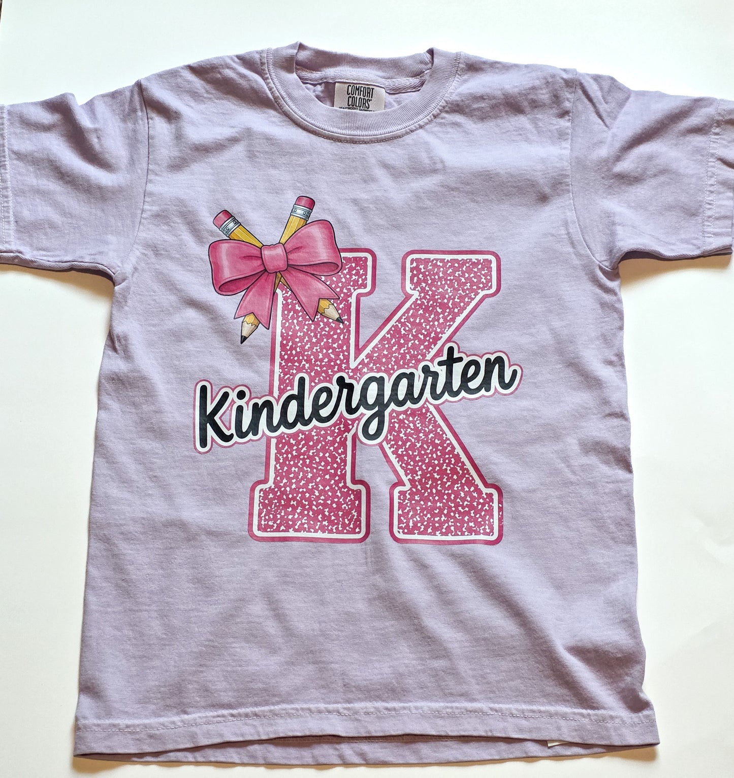 A light lavender-gray short-sleeve t-shirt laid flat on a white background, featuring a graphic design with a large pink glitter-textured letter 'K', a pink bow with two pencils, and the word 'Kindergarten' in black script with a white outline.