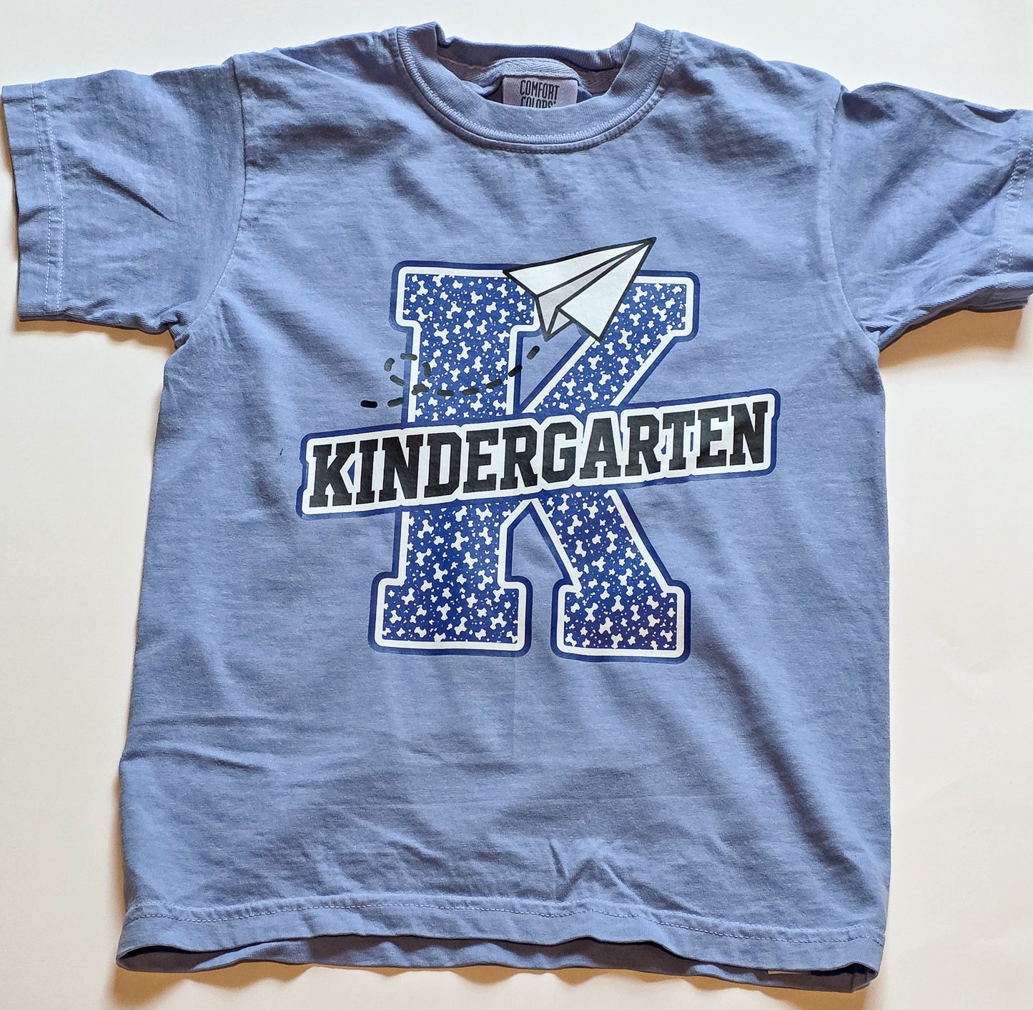 A light blue short-sleeve kids' t-shirt laid flat on a light beige background. The shirt features a graphic design with a large blue star-patterned letter 'K', a white paper airplane, and the word 'KINDERGARTEN' in black and white block letters.