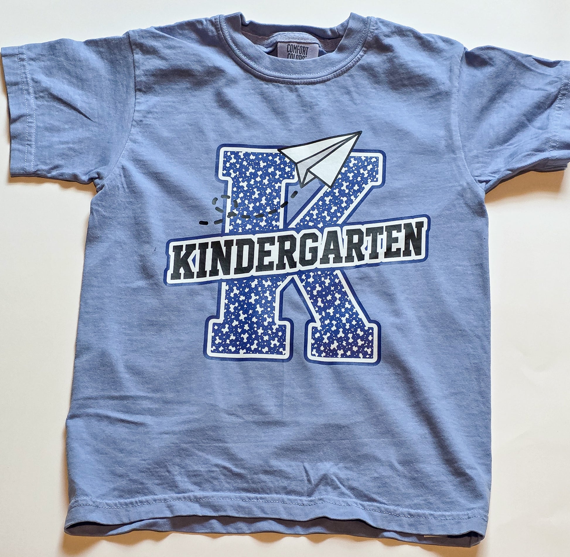 A light blue short-sleeve kids' t-shirt laid flat on a light beige background. The shirt features a graphic design with a large blue star-patterned letter 'K', a white paper airplane, and the word 'KINDERGARTEN' in black and white block letters.