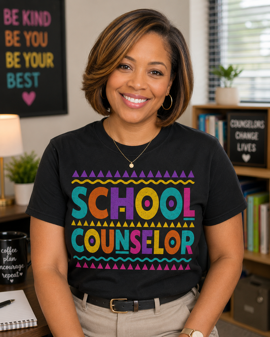 Black woman with a bob and honey blonde highlights wearing a black teacher that says SCHOOL COUNSELOR in bright colors, teal, orange, purple, yellow and hot pink.