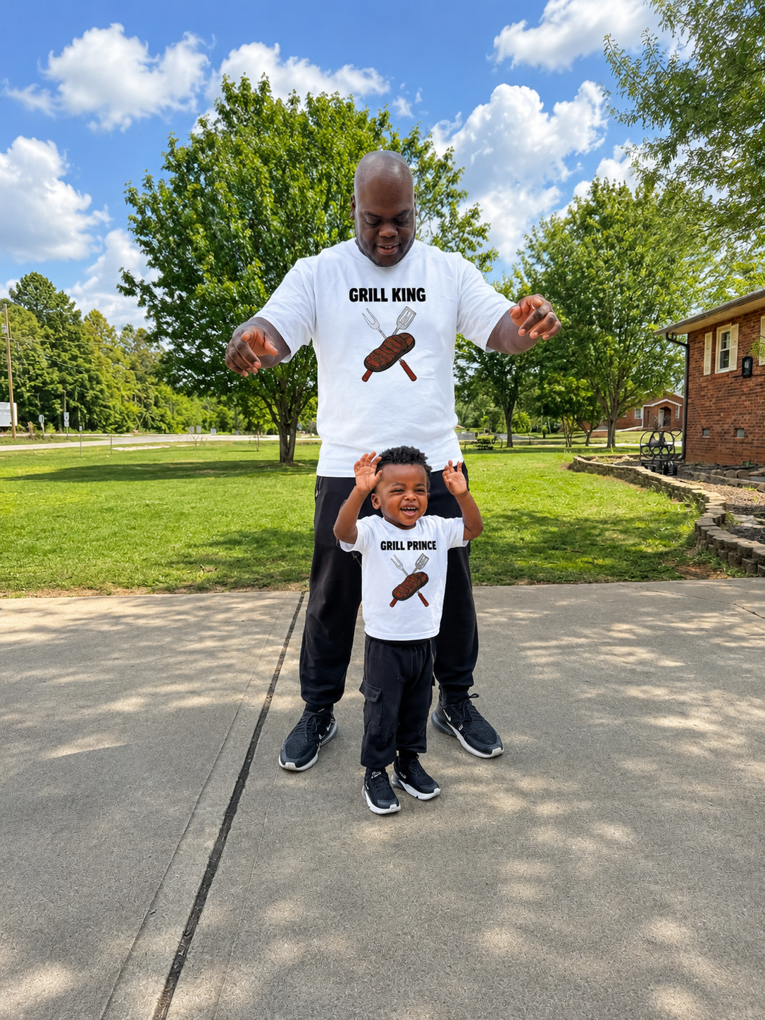 A man and a young boy standing outdoors on a paved path, both wearing matching white graphic T-shirts with a grill design and the words 'GRILL KING' and 'GRILL PRINCE' respectively, against a background of green trees and a blue sky with clouds.
